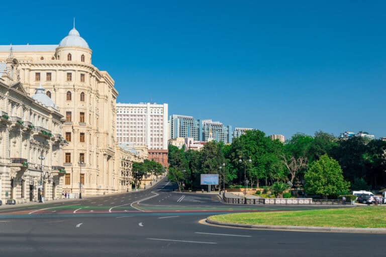 רחוב עיר רחב עם בניינים ישנים בצבע בז' משמאל, בנייני קומות מודרניים ברקע, ואזור פארק ירוק מימין תחת שמיים כחולים בהירים. - Kerato Life מה שלא עשה הטבע עושה הצבע