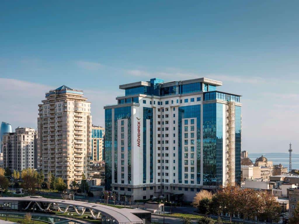 בניין מלון מודרני וגבוה עם חלונות זכוכית ושלט Mövenpick, מוקף בבניינים אחרים ועצים תחת שמיים כחולים וצלולים — נוף מדהים המזכיר את האפקט המהפכני של שינוי צבע עיניים. - Kerato Life מה שלא עשה הטבע עושה הצבע